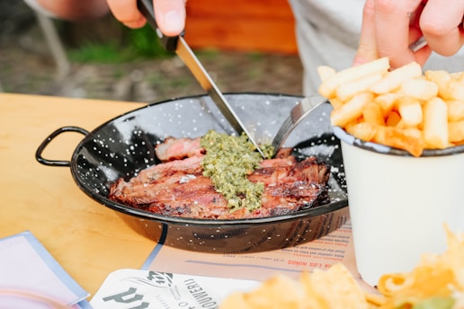 A person is using a knife and fork to cut into a piece of steak topped with a green sauce. The steak is served in a black speckled pan, and there is a cup filled with French fries placed nearby on a wooden table.