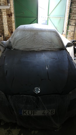 A car is parked inside a garage, covered with a protective plastic sheet. The vehicle is positioned in front of a slightly ajar green door, with a visible license plate labeled 'KUF 426'. Brick walls and a concrete floor surround the scene, under natural and dim lighting.