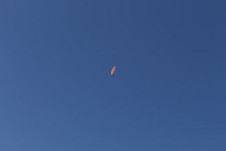 A solitary orange paraglider gliding smoothly through a vast, clear blue sky.