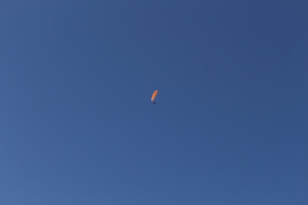 A solitary orange paraglider gliding smoothly through a vast, clear blue sky.