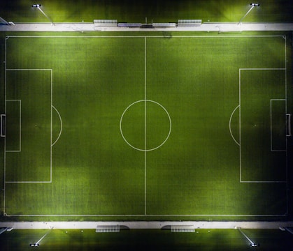An aerial view of a football field at night, illuminated by floodlights. The green field has well-marked white lines for the playing area, including the center circle, penalty boxes, and sidelines. Benches and walkways are visible along the top edge of the image.