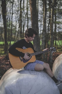 A person is sitting on a large round object in a forested area, playing an acoustic guitar. The individual is wearing a black long-sleeved shirt and light shorts, focused on playing the instrument.