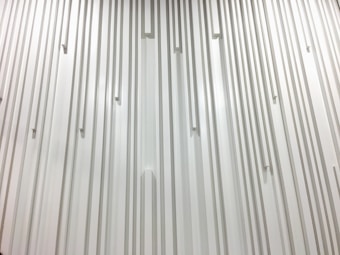 A textured wall featuring vertical lines and rectangular protrusions evenly spaced across the surface, creating a rhythmic pattern. The monochromatic design emphasizes the play of light and shadow on a smooth, minimalist architectural feature.