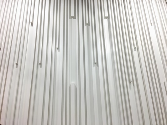 A textured wall featuring vertical lines and rectangular protrusions evenly spaced across the surface, creating a rhythmic pattern. The monochromatic design emphasizes the play of light and shadow on a smooth, minimalist architectural feature.