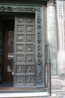 An intricately designed door with detailed carvings, next to a stone pillar. The door features multiple panels, each with unique relief sculptures depicting various scenes and characters. Adjacent to the door are ornate floral decorations and a partially open wrought iron gate. The surrounding architecture includes a column with visible wear, and part of a decorative tiled wall.