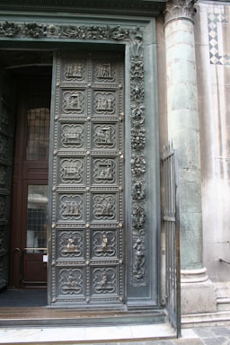 An intricately designed door with detailed carvings, next to a stone pillar. The door features multiple panels, each with unique relief sculptures depicting various scenes and characters. Adjacent to the door are ornate floral decorations and a partially open wrought iron gate. The surrounding architecture includes a column with visible wear, and part of a decorative tiled wall.
