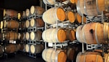 A storage area filled with numerous wooden barrels, stacked in metal racks. The barrels appear to be used for aging wine or other spirits, with varying shades and stains indicating use.
