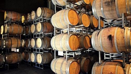 A storage area filled with numerous wooden barrels, stacked in metal racks. The barrels appear to be used for aging wine or other spirits, with varying shades and stains indicating use.