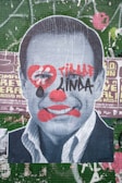 A large poster depicts a black-and-white portrait of a man's face overlaid with graffiti-style art. The face has red clown-like makeup, including exaggerated lips, a nose, and a heart shape around the eyes. Bold red and black graffiti text is also present across the face. The background consists of layers of worn and torn posters on a green tiled wall with various text and graphics.