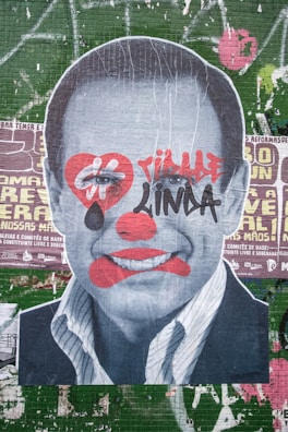 A large poster depicts a black-and-white portrait of a man's face overlaid with graffiti-style art. The face has red clown-like makeup, including exaggerated lips, a nose, and a heart shape around the eyes. Bold red and black graffiti text is also present across the face. The background consists of layers of worn and torn posters on a green tiled wall with various text and graphics.