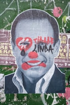 A large poster depicts a black-and-white portrait of a man's face overlaid with graffiti-style art. The face has red clown-like makeup, including exaggerated lips, a nose, and a heart shape around the eyes. Bold red and black graffiti text is also present across the face. The background consists of layers of worn and torn posters on a green tiled wall with various text and graphics.