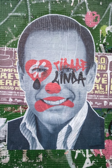 A large poster depicts a black-and-white portrait of a man's face overlaid with graffiti-style art. The face has red clown-like makeup, including exaggerated lips, a nose, and a heart shape around the eyes. Bold red and black graffiti text is also present across the face. The background consists of layers of worn and torn posters on a green tiled wall with various text and graphics.