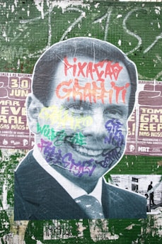 A large street art mural featuring a black-and-white cutout image of a man in a suit. The portrait is covered with colorful graffiti tags in various styles and colors, including red, purple, yellow, and green. The background consists of a green tiled wall with painted over posters and additional graffiti.