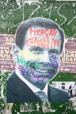 A large street art mural featuring a black-and-white cutout image of a man in a suit. The portrait is covered with colorful graffiti tags in various styles and colors, including red, purple, yellow, and green. The background consists of a green tiled wall with painted over posters and additional graffiti.