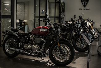 A row of sleek motorcycles is on display in a showroom setting. The motorcycles are primarily black and red, with chrome detailing, and are lined up side by side. The room is dimly lit, creating a dramatic and sophisticated atmosphere.