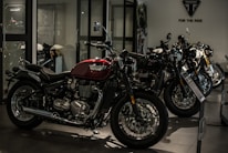 A row of sleek motorcycles is on display in a showroom setting. The motorcycles are primarily black and red, with chrome detailing, and are lined up side by side. The room is dimly lit, creating a dramatic and sophisticated atmosphere.