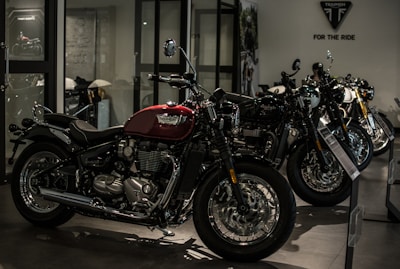 A row of sleek motorcycles is on display in a showroom setting. The motorcycles are primarily black and red, with chrome detailing, and are lined up side by side. The room is dimly lit, creating a dramatic and sophisticated atmosphere.