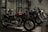 A row of sleek motorcycles is on display in a showroom setting. The motorcycles are primarily black and red, with chrome detailing, and are lined up side by side. The room is dimly lit, creating a dramatic and sophisticated atmosphere.