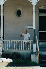 An elderly man with a gray beard and white hair, dressed in white clothing, sits on a wooden porch. He is waving with his right hand and appears to be looking directly ahead. The porch and house behind him have an old, weathered appearance with peeling paint. Decorative woodwork frames the porch, and there is a mailbox attached to the wall near the front door. The house number '121' is partially visible above the door.