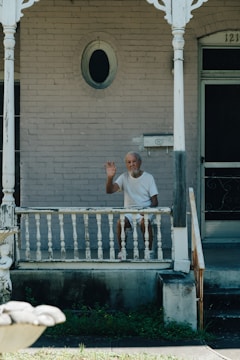 An elderly man with a gray beard and white hair, dressed in white clothing, sits on a wooden porch. He is waving with his right hand and appears to be looking directly ahead. The porch and house behind him have an old, weathered appearance with peeling paint. Decorative woodwork frames the porch, and there is a mailbox attached to the wall near the front door. The house number '121' is partially visible above the door.