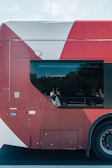 A red and white bus with a large window reflects a person taking a photo. The bus is parked on a street, and trees and a fence are visible in the background. The bus shows some marks and has a CNG label on the side.