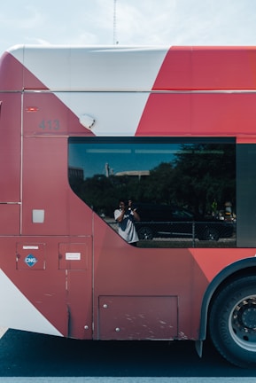A red and white bus with a large window reflects a person taking a photo. The bus is parked on a street, and trees and a fence are visible in the background. The bus shows some marks and has a CNG label on the side.