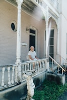 An elderly person is sitting on a chair at the porch of an older wooden house with peeling paint. The porch has intricate, vintage-style railings and columns. The house is painted in a light color, with some damage visible on the wood. In the foreground, there is a worn-out fountain or birdbath surrounded by some grass.