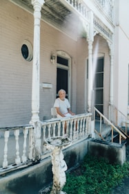 An elderly person is sitting on a chair at the porch of an older wooden house with peeling paint. The porch has intricate, vintage-style railings and columns. The house is painted in a light color, with some damage visible on the wood. In the foreground, there is a worn-out fountain or birdbath surrounded by some grass.
