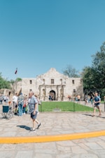 People walking and gathering in front of an old historic building with a rustic facade. A Texas flag is visible on the left, and trees flank the structure. The plaza area has stone paving and a boundary of yellow curb.