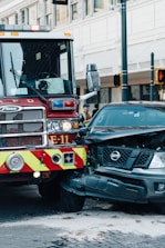 A fire truck and a black Nissan vehicle are involved in a collision at an urban intersection. The front of the car is severely damaged and white powder is visible on the ground, possibly from a fire extinguisher. The surroundings include city buildings and traffic lights.