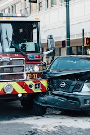 A fire truck and a black Nissan vehicle are involved in a collision at an urban intersection. The front of the car is severely damaged and white powder is visible on the ground, possibly from a fire extinguisher. The surroundings include city buildings and traffic lights.