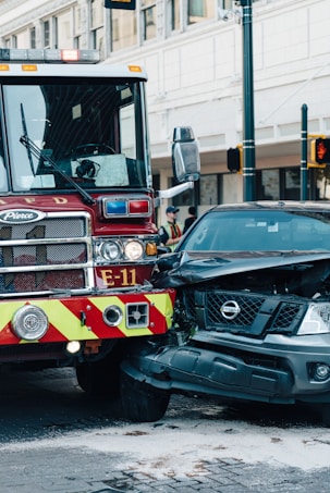 A fire truck and a black Nissan vehicle are involved in a collision at an urban intersection. The front of the car is severely damaged and white powder is visible on the ground, possibly from a fire extinguisher. The surroundings include city buildings and traffic lights.