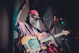 A musician with a long white beard and wearing a red headscarf is passionately performing on stage. He is playing an electric bass guitar and is in mid-motion, suggesting a lively or intense performance. The lighting is dim with a focus on the musician, creating an intimate atmosphere.