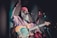 A musician with a long white beard and wearing a red headscarf is passionately performing on stage. He is playing an electric bass guitar and is in mid-motion, suggesting a lively or intense performance. The lighting is dim with a focus on the musician, creating an intimate atmosphere.