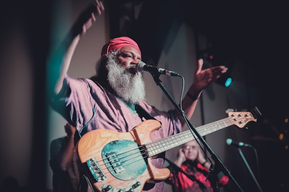A musician with a long white beard and wearing a red headscarf is passionately performing on stage. He is playing an electric bass guitar and is in mid-motion, suggesting a lively or intense performance. The lighting is dim with a focus on the musician, creating an intimate atmosphere.