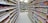A well-organized supermarket aisle with shelves stocked with a variety of food products, such as condiments, spices, instant foods, and convenience items. The shelves are fully stocked and well-lit, displaying a wide range of colorful packaging. At the end of the aisle, potted plants and flowers are visible, adding a touch of greenery and freshness.