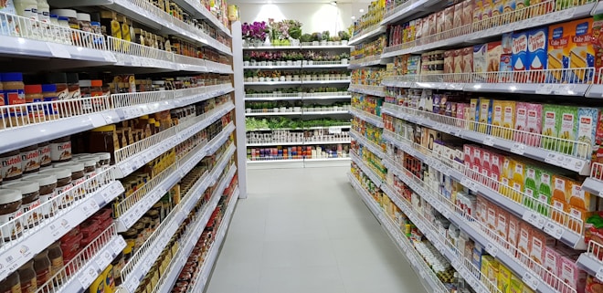 A well-organized supermarket aisle with shelves stocked with a variety of food products, such as condiments, spices, instant foods, and convenience items. The shelves are fully stocked and well-lit, displaying a wide range of colorful packaging. At the end of the aisle, potted plants and flowers are visible, adding a touch of greenery and freshness.