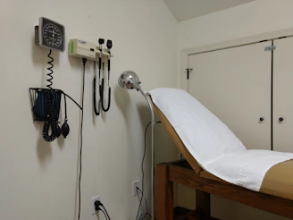 A medical examination room featuring an examination table covered with a white paper sheet. Medical instruments such as a blood pressure gauge, otoscope, and ophthalmoscope are mounted on the wall. The furnishings include light-colored walls and cabinets.