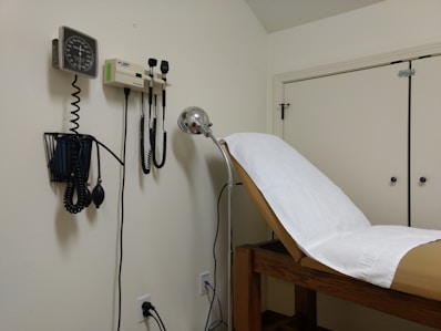 A medical examination room featuring an examination table covered with a white paper sheet. Medical instruments such as a blood pressure gauge, otoscope, and ophthalmoscope are mounted on the wall. The furnishings include light-colored walls and cabinets.