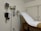 A medical examination room featuring an examination table covered with a white paper sheet. Medical instruments such as a blood pressure gauge, otoscope, and ophthalmoscope are mounted on the wall. The furnishings include light-colored walls and cabinets.