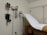 A medical examination room featuring an examination table covered with a white paper sheet. Medical instruments such as a blood pressure gauge, otoscope, and ophthalmoscope are mounted on the wall. The furnishings include light-colored walls and cabinets.