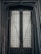 A close-up view of a black metal door or window with a grid pattern, featuring frosted or translucent glass panes behind the grid. The surrounding frame appears worn, with signs of weathering.