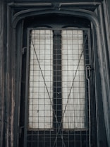A close-up view of a black metal door or window with a grid pattern, featuring frosted or translucent glass panes behind the grid. The surrounding frame appears worn, with signs of weathering.