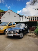 A classic black car with a vintage design is parked in a driveway next to a modern, yellow car. The background features a row of houses, with white exteriors and brown roof tiles. The sky is mostly blue, speckled with a few white clouds.