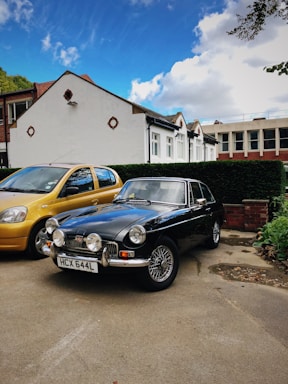 A classic black car with a vintage design is parked in a driveway next to a modern, yellow car. The background features a row of houses, with white exteriors and brown roof tiles. The sky is mostly blue, speckled with a few white clouds.
