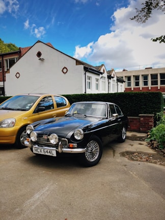 A classic black car with a vintage design is parked in a driveway next to a modern, yellow car. The background features a row of houses, with white exteriors and brown roof tiles. The sky is mostly blue, speckled with a few white clouds.