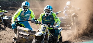 A group of motocross racers wearing bright blue and yellow gear is participating in a sidecar motocross race. The vehicles kick up clouds of dirt as they speed around a track. The riders wear helmets and goggles for protection. The scene is dynamic and action-packed with dust creating an intense atmosphere.
