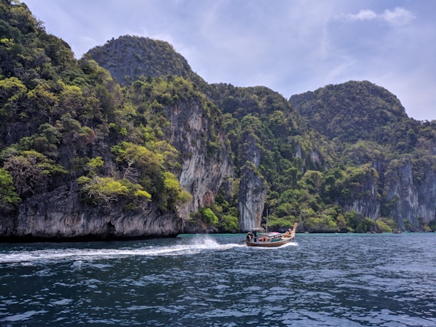 A serene coastal scene with lush green cliffs rising dramatically from the deep blue sea. A traditional long-tail boat moves swiftly across the water, leaving a trail of white foam.