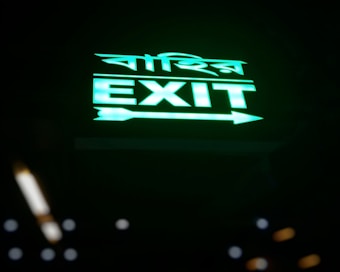 A green illuminated exit sign is prominently displayed, showing the word 'EXIT' in bold letters along with another script, possibly a different language. The sign features an arrow pointing right, and the background is dark to highlight the glow of the text.