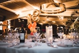 A dimly lit restaurant or event space with a large gold balloon shaped like the number 18 is surrounded by clusters of smaller orange and gold balloons. There are elegantly set tables with wine glasses, cutlery, and a bottle of wine. The ceiling features modern architectural elements and lighting, giving a warm ambiance.